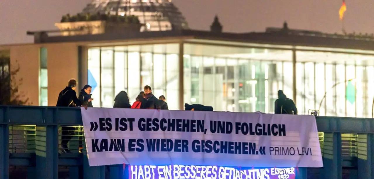 Man erkennt eine Brücke im Vordergrund, an der Personen ein Banner mit der Aufschrift "Es ist geschehen, und folglich kann es wieder geschehen." (Primo Levi) angebracht haben. Im Hintergrund ist unscharf das Reichstagsgebäude mit der Glaskuppel zu erkennen.