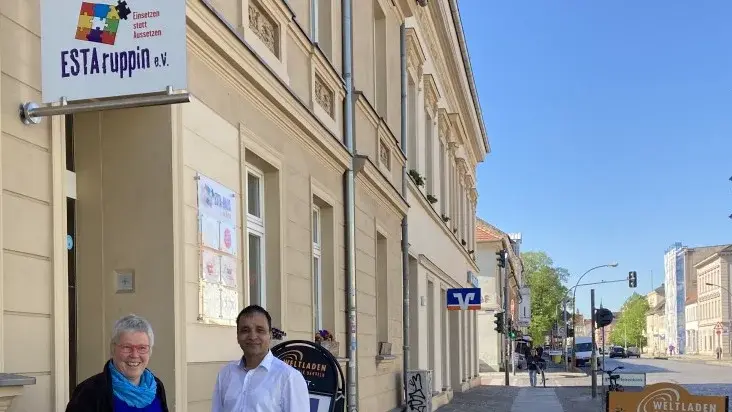 Links ist das ESTA-haus der Vielfalt in Neuruppin zu sehen, vor der Eingangstür stehen Pfarrerin Christiane Schulz, Geschäftsführerin von ESTAruppin e. V. und ESTAruppin-Mitarbeiter Ferdydun Rahimi in der Bildmitte.
