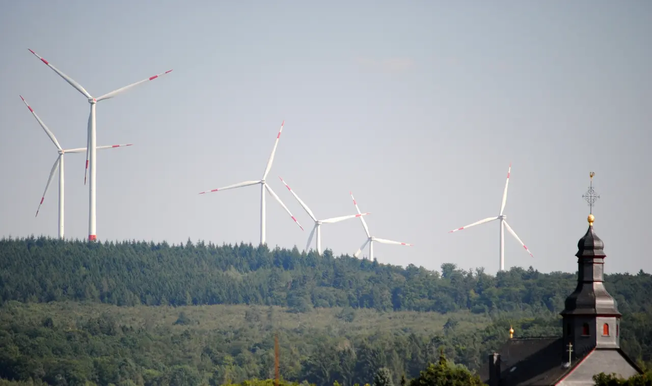 Auf dem Bild sind im Hintergrund mehrere Windräder zu sehen, welche sich vom Himmel abheben. Im Vordergrund sieht man einen Kirchturm.