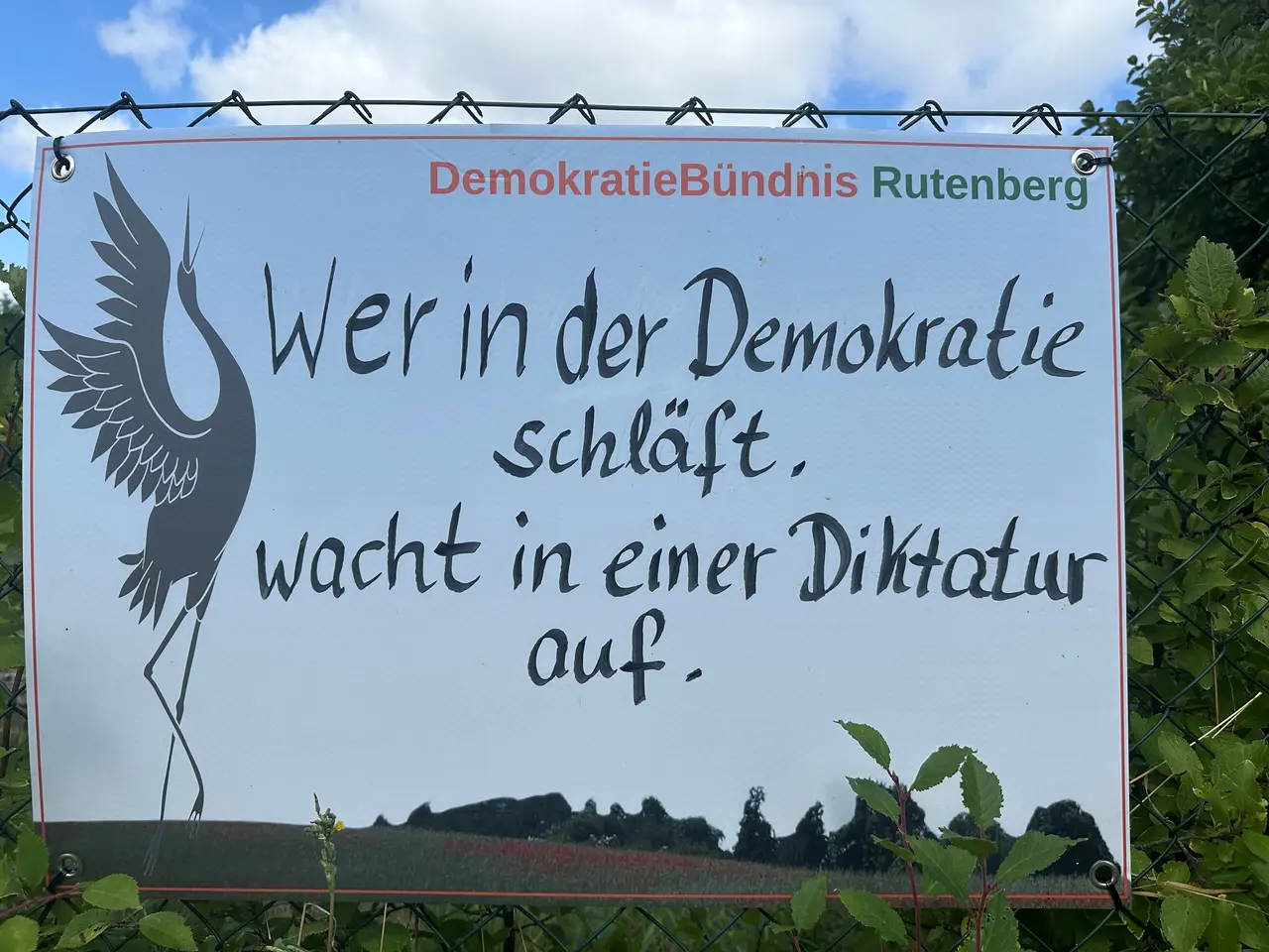 Man erkennt ein Banner an einem Maschendrahtzaun. Die Aufschrift zeigt das Logo des DemokratieBündnisses Rutenberg e. V. einen Kranich mit dem Text &quot;Wer in der Demokratie schläft, wacht in einer Diktatur auf.&quot;