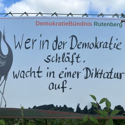 Man erkennt ein Banner an einem Maschendrahtzaun. Die Aufschrift zeigt das Logo des DemokratieBündnisses Rutenberg e. V. einen Kranich mit dem Text "Wer in der Demokratie schläft, wacht in einer Diktatur auf."