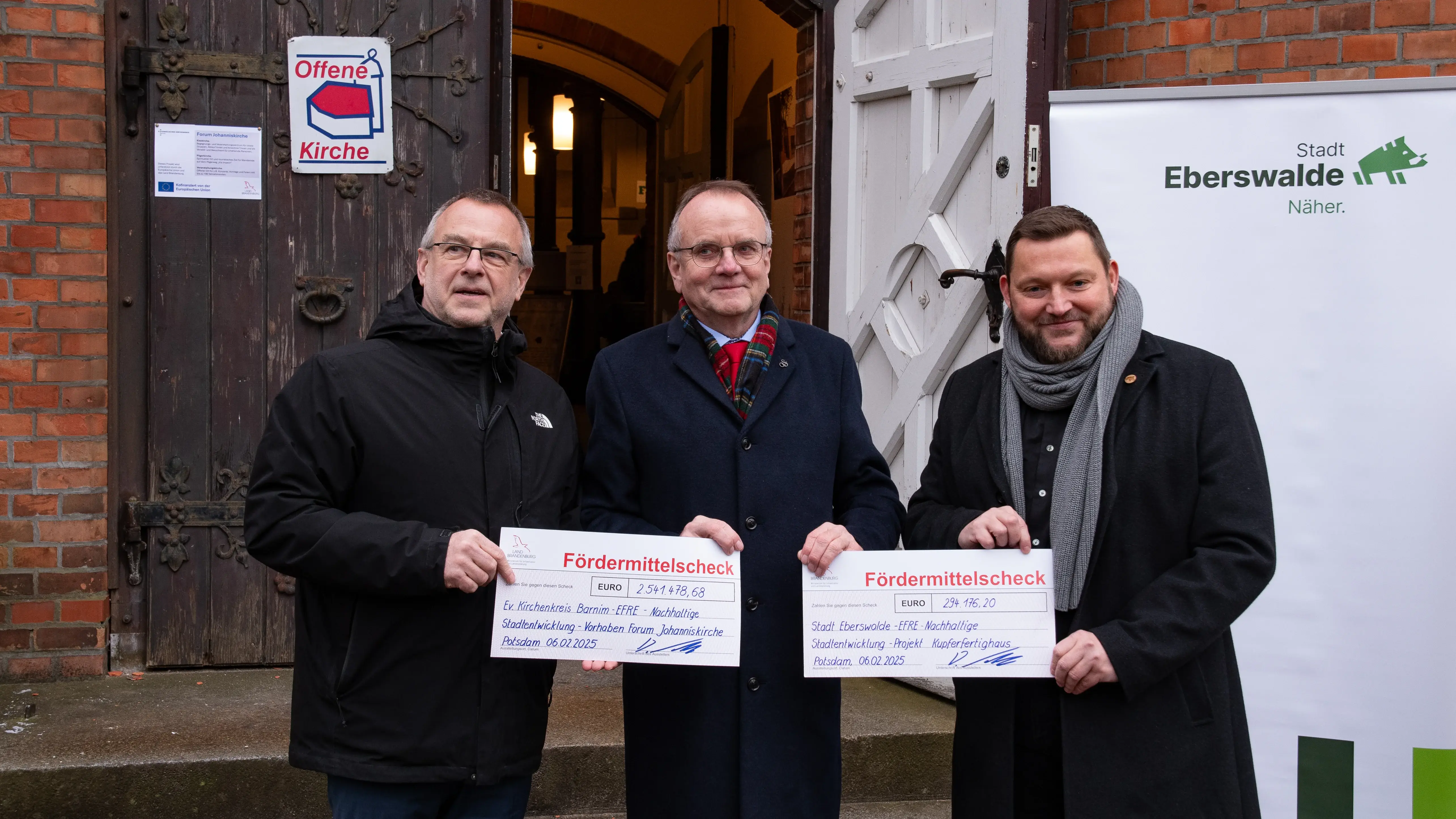 Man erkennt der Minister für Infrastruktur und Landesplanung des Landes Brandenburg, Detlef Tabbert (Mitte des Bildes), vor der geöffenten Kirchentür der Johanniskirche. Er übergibt Fördermittelschecks an Pfarrer Christoph Brust (links im Bild) und Bürgermeister Götz Herrmann (rechts im Bild).