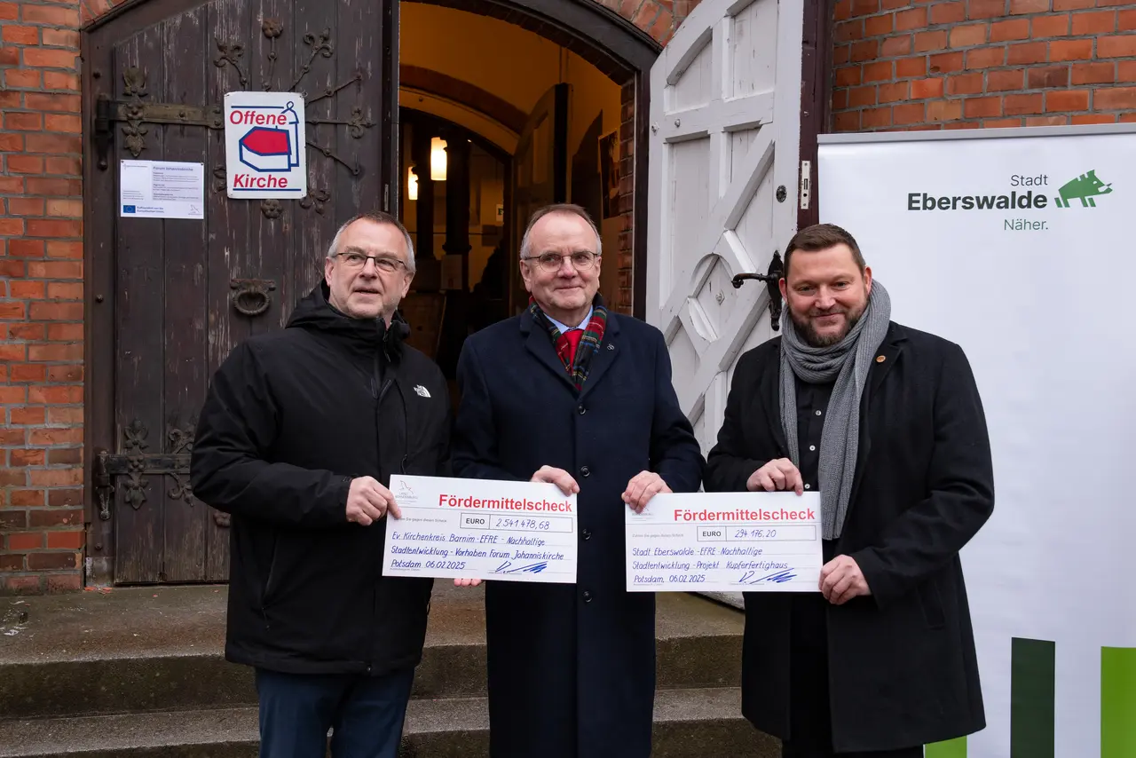 Man erkennt der Minister für Infrastruktur und Landesplanung des Landes Brandenburg, Detlef Tabbert (Mitte des Bildes), vor der geöffenten Kirchentür der Johanniskirche. Er übergibt Fördermittelschecks an Pfarrer Christoph Brust (links im Bild) und Bürgermeister Götz Herrmann (rechts im Bild).