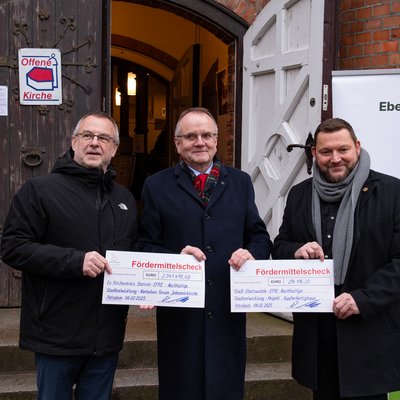 Man erkennt der Minister für Infrastruktur und Landesplanung des Landes Brandenburg, Detlef Tabbert (Mitte des Bildes), vor der geöffenten Kirchentür der Johanniskirche. Er übergibt Fördermittelschecks an Pfarrer Christoph Brust (links im Bild) und Bürgermeister Götz Herrmann (rechts im Bild).
