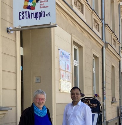 Links ist das ESTA-haus der Vielfalt in Neuruppin zu sehen, vor der Eingangstür stehen Pfarrerin Christiane Schulz, Geschäftsführerin von ESTAruppin e. V. und ESTAruppin-Mitarbeiter Ferdydun Rahimi in der Bildmitte.